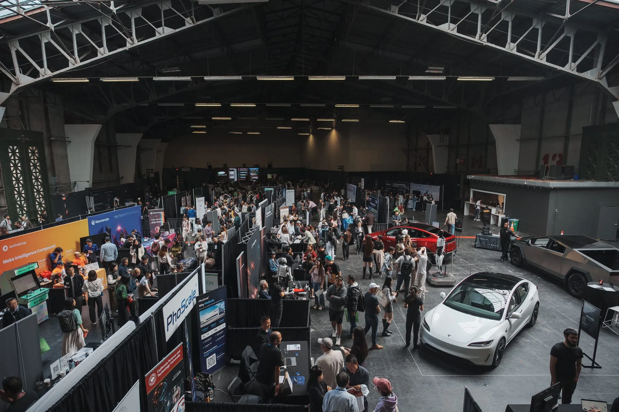 A tech expo floor in full swing seen from above — booths, crowds, and demo cars under an industrial roof, San Francisco