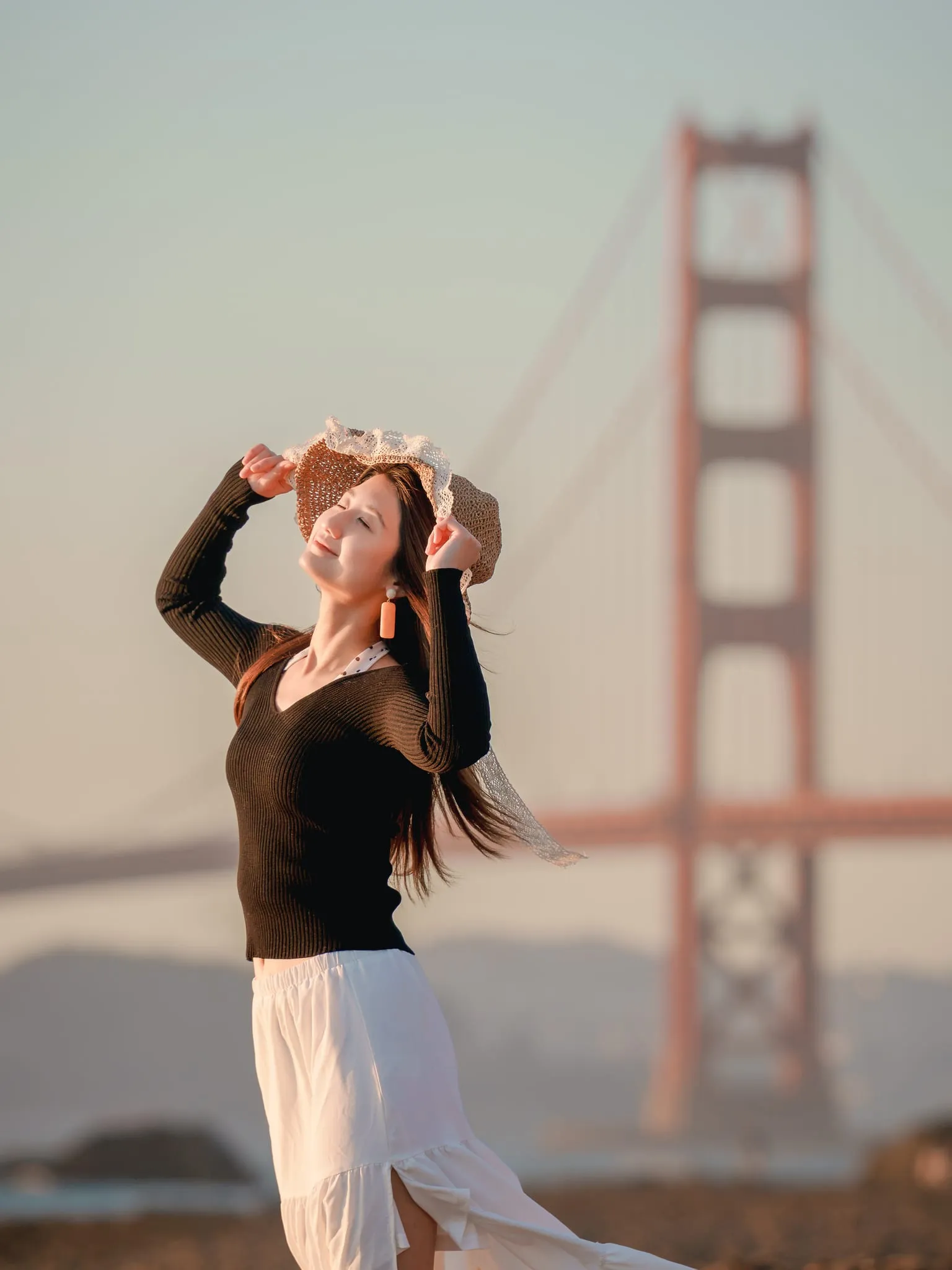Woman in a straw hat basking in golden-hour light with eyes closed, the Golden Gate Bridge soft in the background