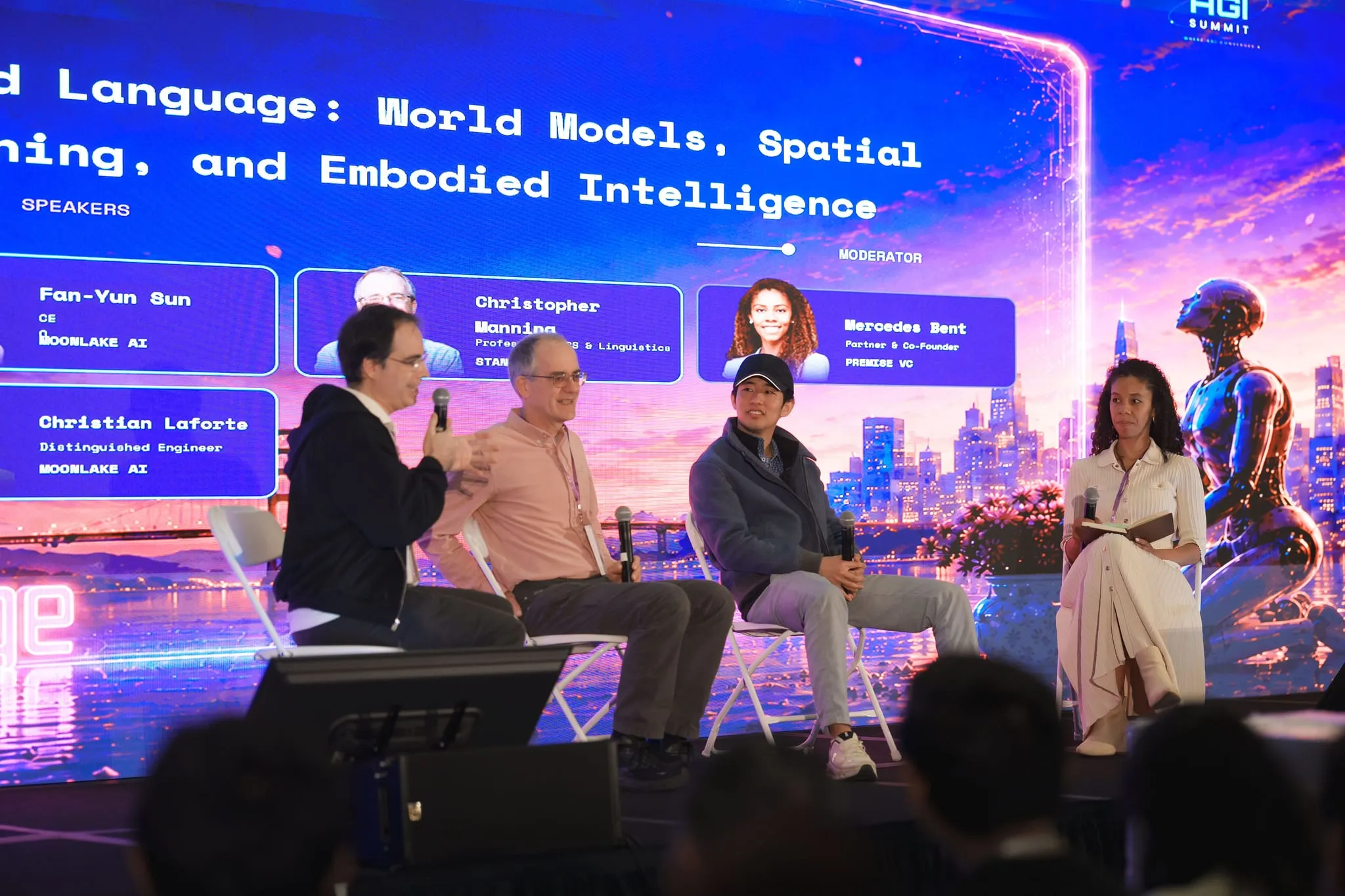 Four panelists in conversation on a summit stage, names on the LED backdrop behind them
