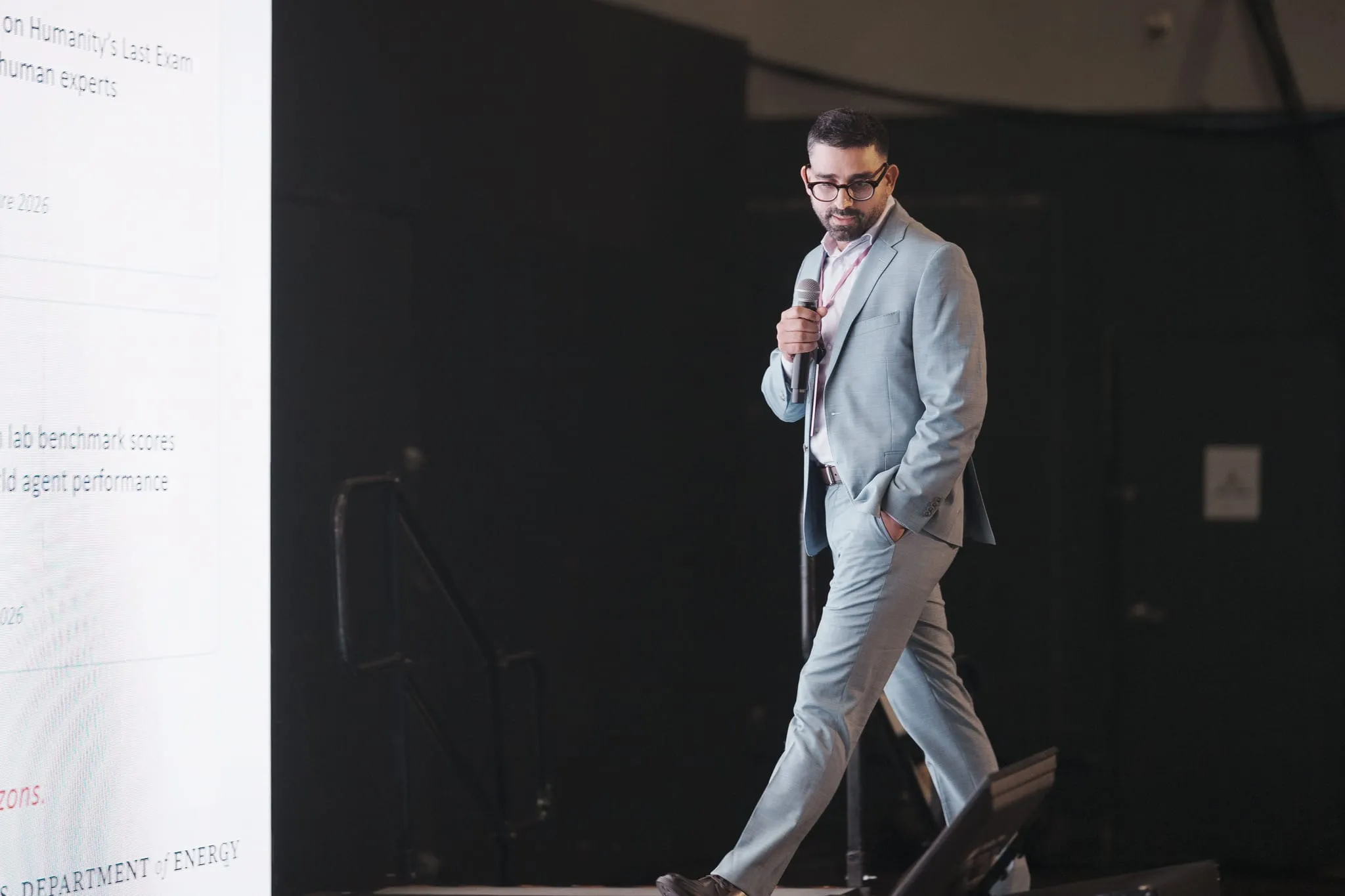 A keynote speaker mid-stride on a dark stage beside a bright slide