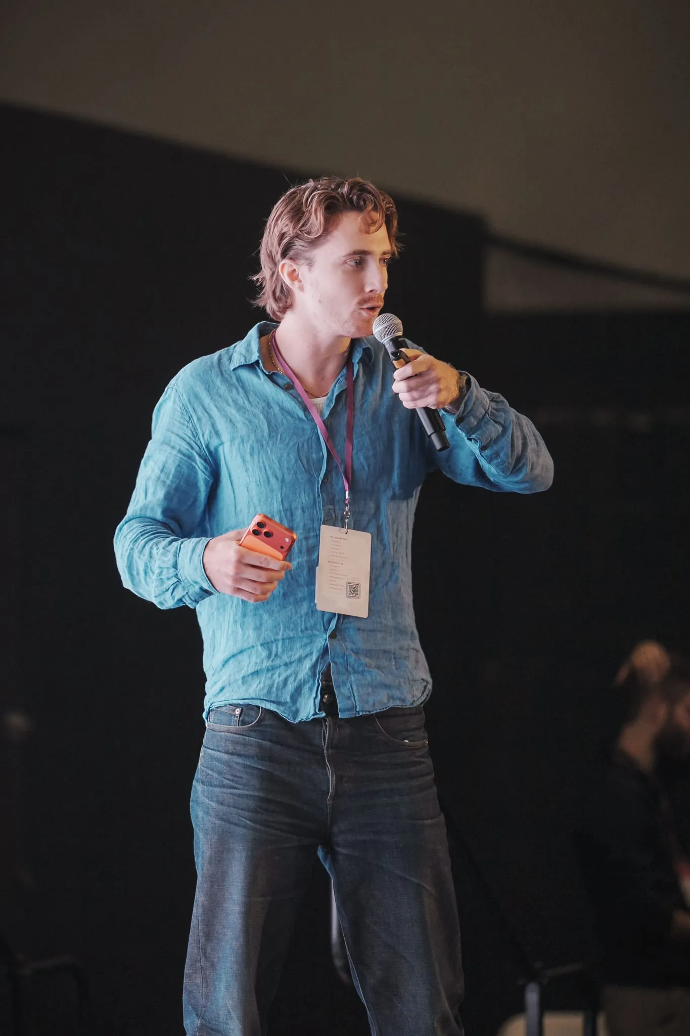 A young founder pitches from the stage, phone in hand, badge swinging